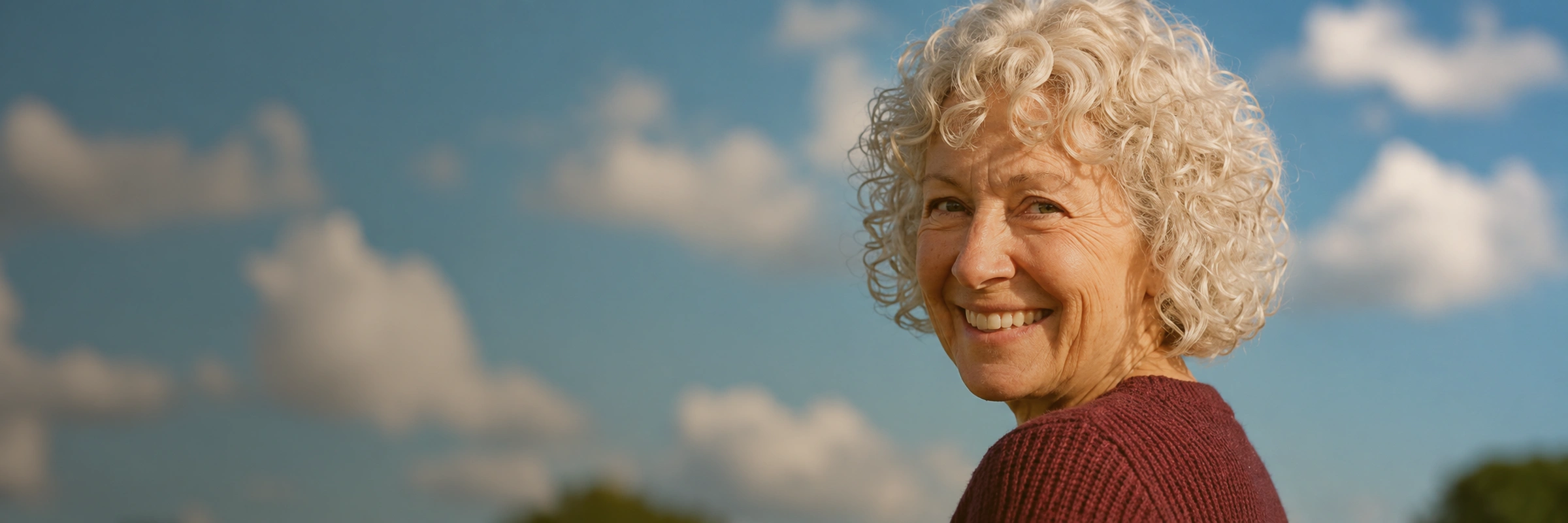 A smiling older woman with curly grey hair outdoors on a bright day